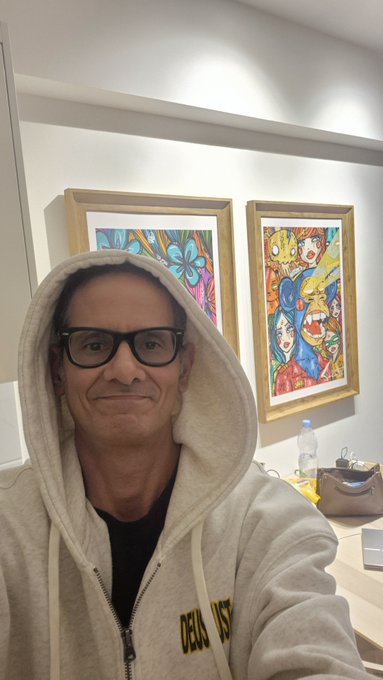 A selfie of a man wearing glasses and a beige hoodie with the hood up, standing in front of two colorful framed paintings in what appears to be a gallery or exhibition space.