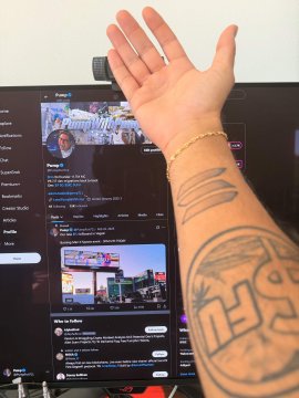 An arm with 'HSR' and other tattoos reaches toward a computer monitor displaying Trump's Truth Social page, with a hand hovering over the screen.