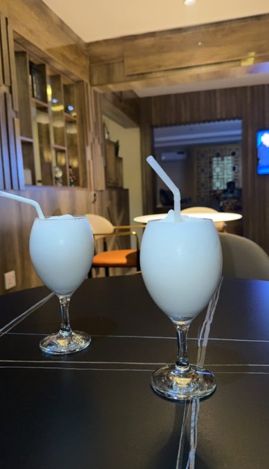 Two glasses of milky white beverages with straws sit on a dark table in what looks like a restaurant or bar interior with wooden accents and warm lighting.