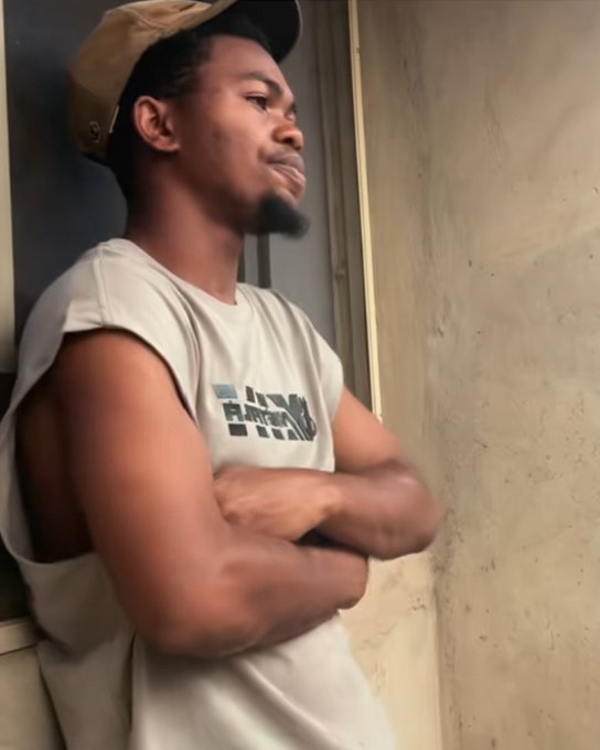 A young man wearing a beige cap and a sleeveless shirt with "RAW" text leans against a wall with his arms crossed, displaying a contemplative or unimpressed expression. His serious, skeptical demeanor contrasts humorously with the post claiming he earned just $2 from a "Grass airdrop," suggesting frustration or disappointment over the meager reward.