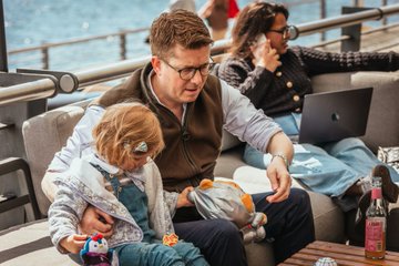 A man wearing glasses and a vest sitting on a couch with a child, working on an Apple laptop with another person in the background.