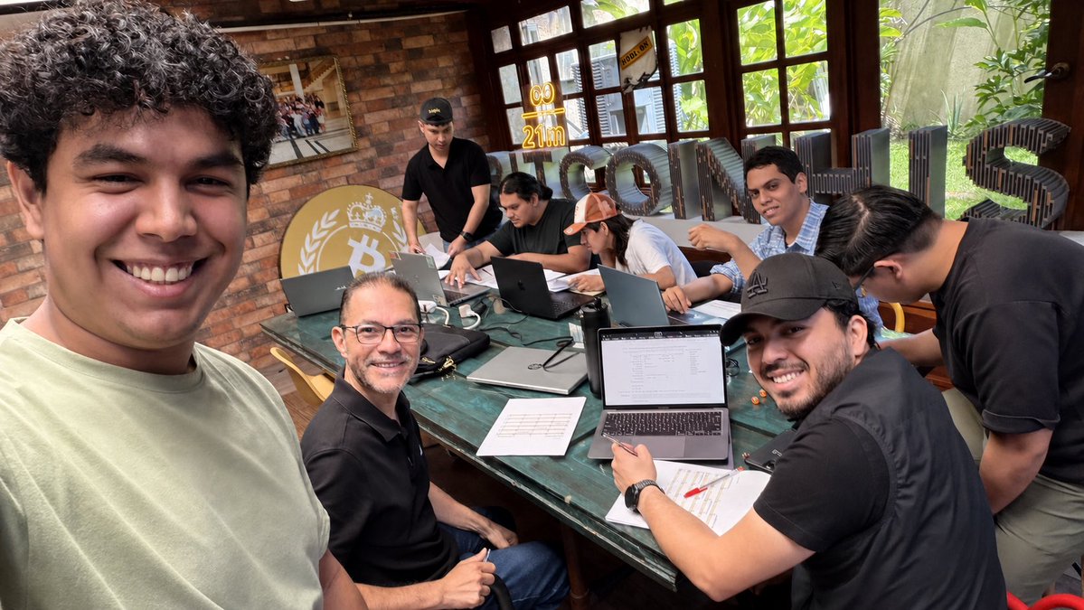 A group of young men smile for a selfie around a green table covered in laptops, with a Bitcoin logo and 'COIN' visible in the background.