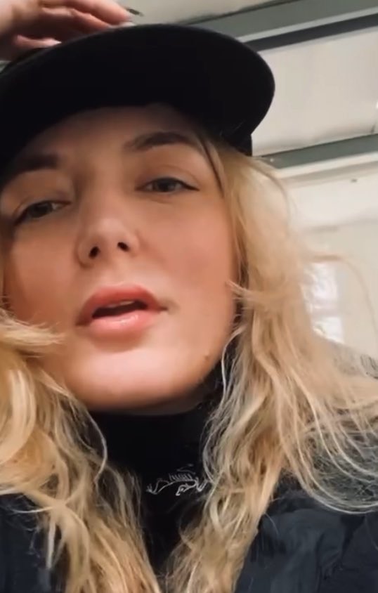 A close-up selfie-style video frame shows a blonde woman wearing a black cap and black t-shirt indoors.