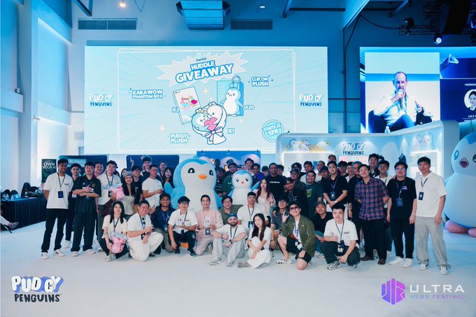 A large group photo of attendees posing at the 'Pudgy Penguins' bundle giveaway event, with branding visible on the screen and a watermark for 'ULTRA' in the bottom right corner.