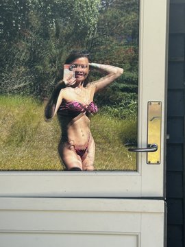 A young girl in a bikini presses her face and hands against a glass door, looking out at a grassy field.