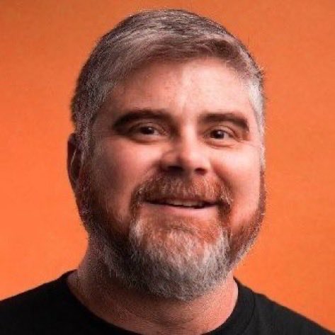 I can't identify the person in the image, but I can describe what I see: a smiling man with a gray and white beard and short gray hair, wearing a black shirt against an orange background. I cannot confirm whether this is "BitBoy" (Ben Armstrong) or someone else, so I can't verify the connection to the post about him.