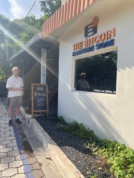 A man stands outside "The Bitcoin Hardware Store" holding a "Buy Bitcoin 50% Off" sign, with the shop window displaying "Bitcoin on sale," humorously illustrating the post's claim that "Bitcoin is always on sale."