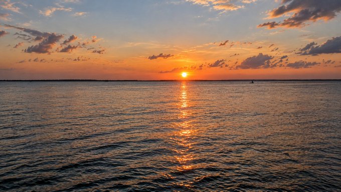 A serene photograph of a sunset over the ocean, with the sun's reflection forming a golden path across the water under a sky filled with scattered clouds.