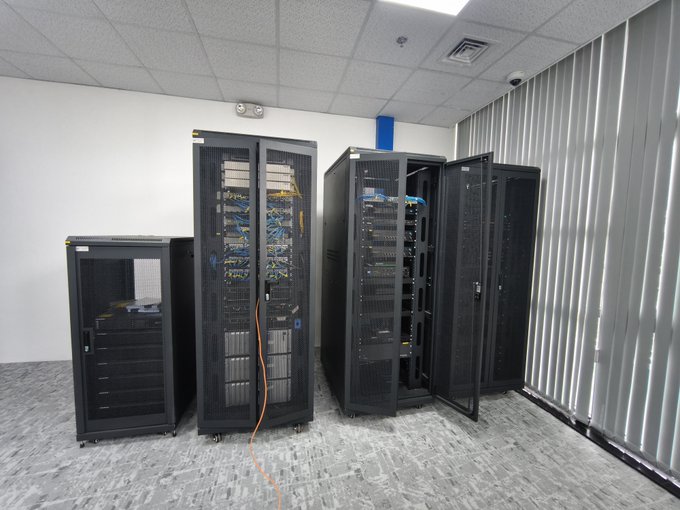 A data center room containing four black server racks with network cabling visible, one rack door open, set against white walls with vertical blinds.