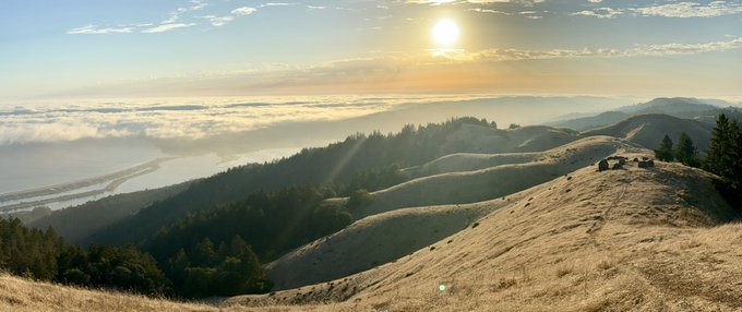 Golden sunlight pours over rolling coastal hills and a sea of low clouds, evoking the poetic image of God "making the clouds a chariot" as the mist drifts above the landscape like a heavenly raft gliding through the sky.