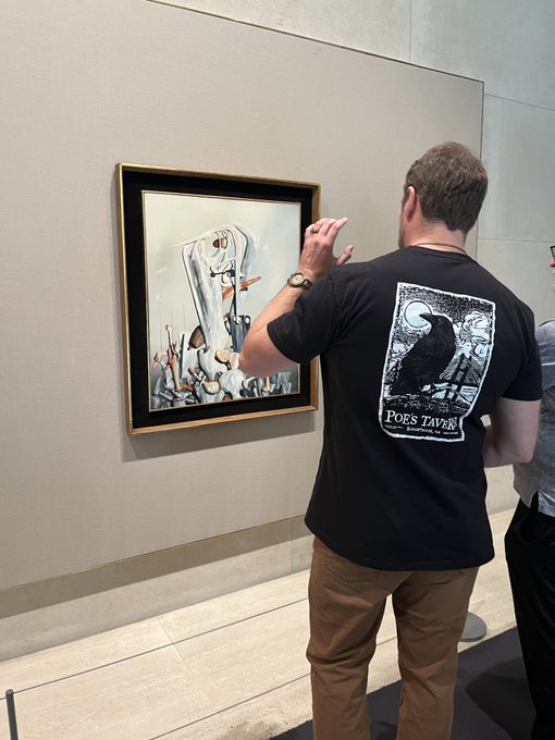 A man wearing a black "Poe's Tavern" t-shirt with a raven graphic stands in a gallery or museum, pointing at a framed surrealist artwork depicting a melting figure.