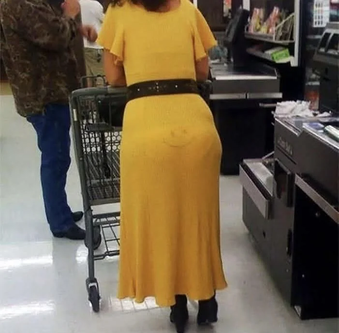 A woman in a yellow dress stands at a grocery store checkout with a smiley face drawn on her backside, viewed from behind as she waits in line.