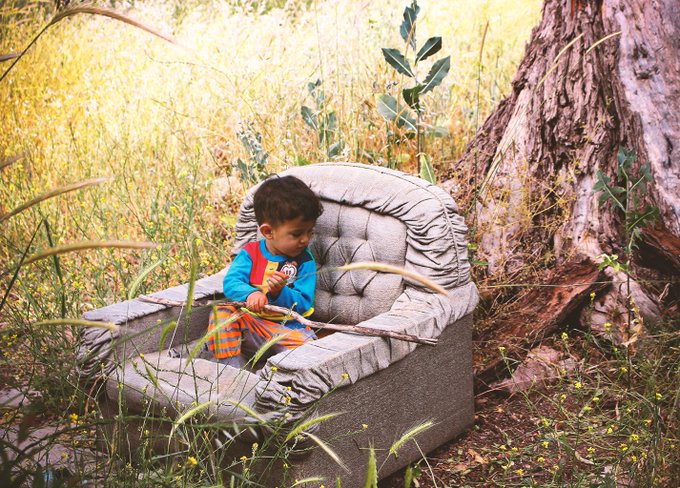 A young boy in a blue shirt and orange-striped overalls sitting in a worn, oversized grey armchair placed outdoors in a grassy field next to a tree.