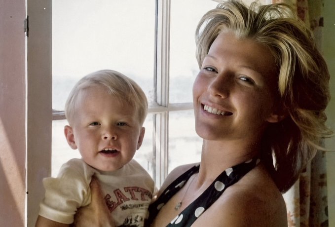 A young blonde woman smiles warmly while holding a smiling toddler in a white "EAT" shirt by a window, capturing a tender mother-and-child moment often associated with young Elon Musk and his mother Maye Musk. The image reflects a nostalgic birthday tribute to Musk's early years.