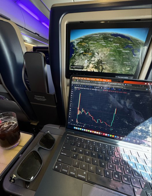 A view from an airplane window shows a flight map, while a laptop displays a red and green stock chart on the tray table alongside sunglasses and a drink.