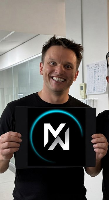 A smiling man with short brown hair wearing a black t-shirt holds up a black square card displaying a stylized white 'M' logo with a glowing cyan ring around it.