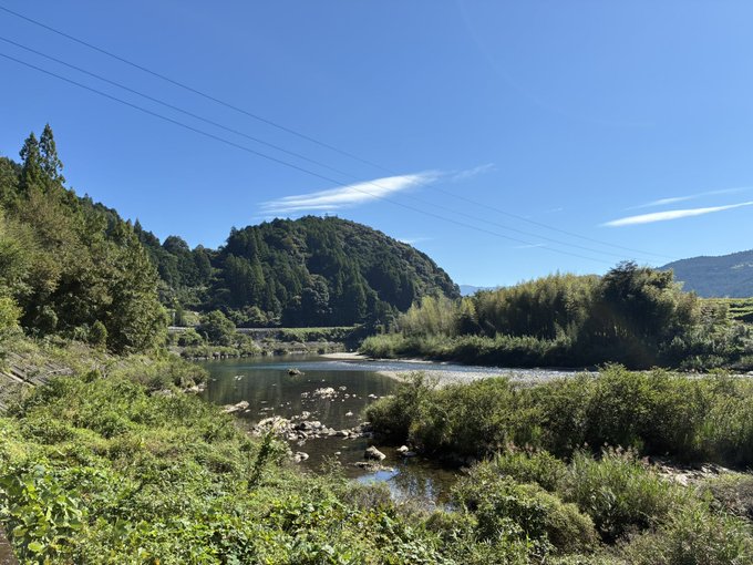 A scenic landscape shows a calm river winding through a rocky bed, flanked by lush green vegetation with forested hills and power lines under a clear blue sky.