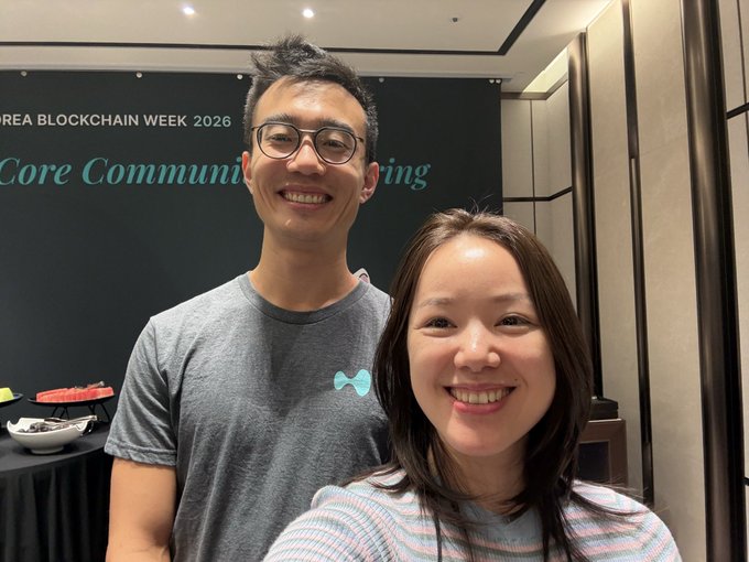 A selfie of a smiling young man and woman at an event, with a backdrop partially visible reading '...REA BLOCKCHAIN WEEK 2026' and '...Core Communit... Meeting'.