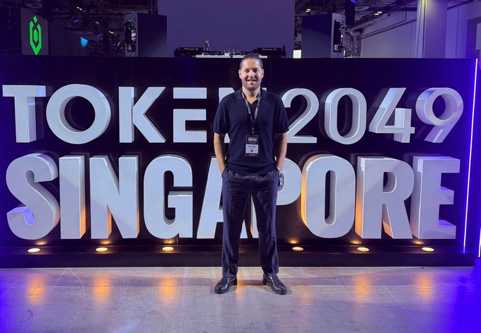 A man wearing a black polo shirt and a lanyard stands smiling in front of a large illuminated 'TOKEN2049 SINGAPORE' sign.