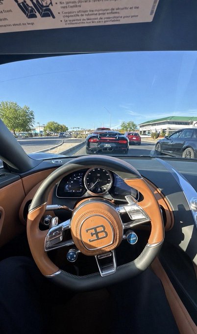 The image shows a view from the driver's seat of a Bugatti, featuring the iconic "BB" logo on the steering wheel, with a Chevrolet Camaro visible ahead through the windshield and a sign partially visible in French at the top. The caption ties to the "gm gm gm" post by highlighting the irony of a rare luxury Bugatti sharing the road with an everyday GM vehicle (the Camaro) at a stoplight.