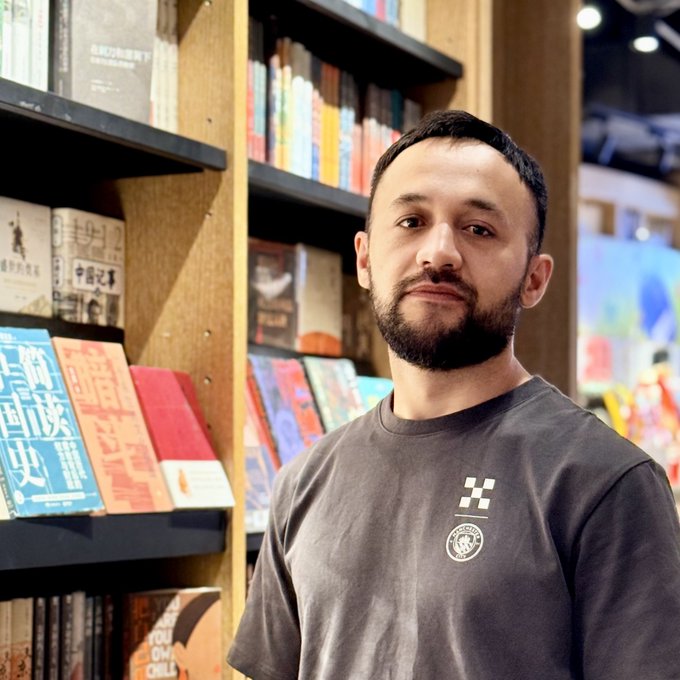 A man with a beard wearing a gray Manchester City t-shirt standing in front of a bookshelf filled with books.