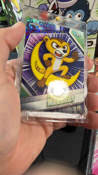 A hand holding a holographic Pokémon trading card featuring an angry-looking creature (appears to be Teddiursa) against a background of colorful Pokémon merchandise.