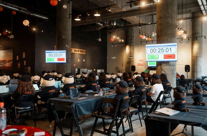 A large room filled with people wearing matching black outfits and hats seated at tables with laptops, with two screens showing '00:25:01' and a 'OUR RIDE' banner visible.