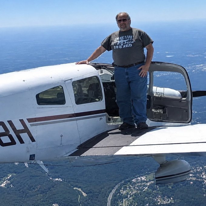 A man wearing a shirt reading "I AM NOT AFRAID FLYING I JUST NOT F**KING IB**ND" stands confidently on the wing of a small white plane with its door open, soaring high above the ground below. The image humorously matches the caption's sentiment—embracing a risk that would terrify most people, just like diving into volatile crypto investments.