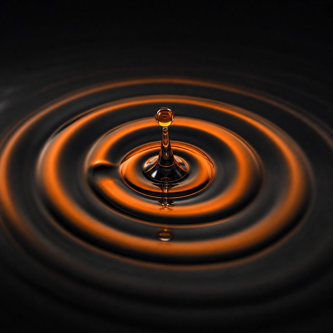 A high-speed photograph of a golden water droplet splashing on a dark liquid surface, creating concentric circular ripples.