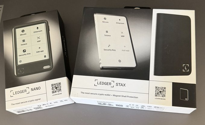 A photograph of two Ledger hardware wallet boxes, the 'LEDGER NANO' and 'LEDGER STAX', both marketed as 'The most secure crypto signer' and 'The most secure crypto wallet • Magnet Shell Protection', supporting Bitcoin, Ethereum, Solana, and XRP.