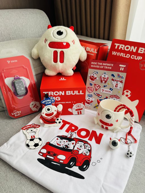 A collection of "TRON BULL" World Cup merchandise is displayed, including a plush toy, ceramic mug, t-shirt, stickers, keychain, and "Lucky Bull" device, all featuring the red bull mascot in a soccer theme. The items relate to the post about receiving a TRON Bull World Cup welcome gift, with the plush being the centerpiece of the themed merchandise bundle.