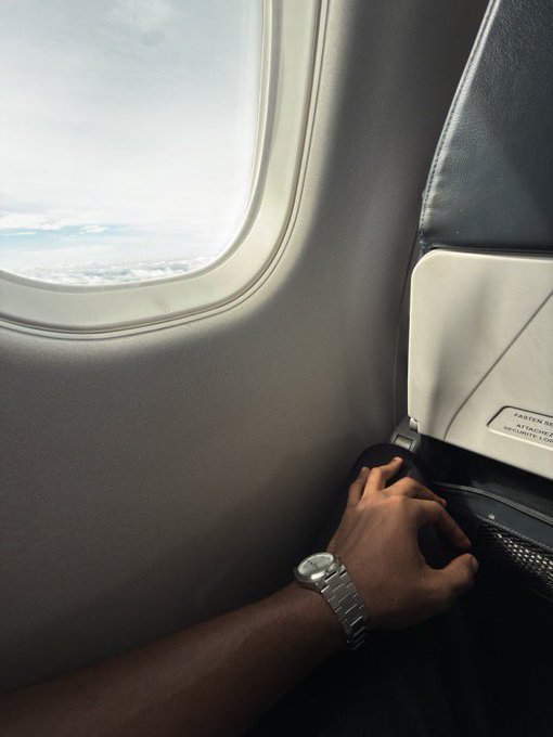 A person's arm wearing a silver wristwatch rests on an airplane tray table by the window, with clouds visible in the sky beyond. A partial label on the seat reads "EXTEN[SION]," "ATTEN[DANCE]," and "[SEAT]BELTS COM[ING]," indicating safety instructions.