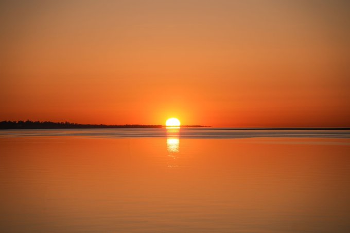 A serene sunset over a calm body of water with the sun reflecting on the surface in vibrant orange hues.