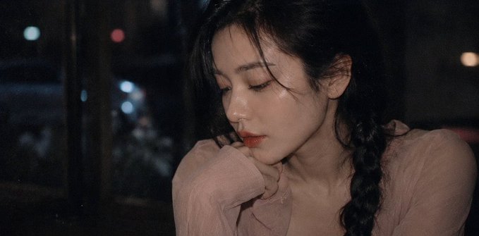 A young woman with long dark braided hair rests her chin on her hands, gazing downward with a contemplative, melancholic expression against a dark background with blurred city lights. The introspective mood of the portrait mirrors the post's reflective tone of ending the day with emotions and wishing followers positivity and beautiful moments ahead.