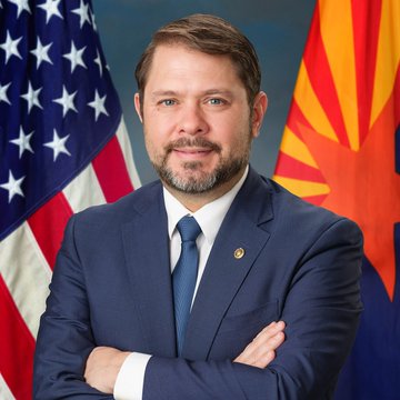 I'm not able to identify the person in the image based on their facial features. I can describe what I see: a man with a beard wearing a dark suit and tie, standing in front of an American flag and the Arizona state flag, suggesting he is an Arizona-based official. A small pin is visible on his lapel.

If this image is being used to represent Senator Ruben Gallego of Arizona in the accompanying post, the Arizona state flag in the background would be contextually relevant, as Gallego represents that state. However, I cannot confirm the identity of the individual shown from their appearance alone. If you can confirm who is pictured, I'd be happy to help with a caption that reflects that context.