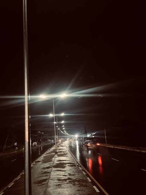 A nighttime view down a wet, reflective road lined with bright streetlights that create prominent lens flares against the dark sky.