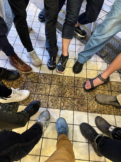 The image shows a group of people standing together, with their feet positioned on a tiled floor. The photo appears to be taken from an overhead perspective, capturing the entire scene. There are at least nine visible individuals in the picture, each wearing different types of shoes and pants. Some of them have their legs crossed or bent, while others stand upright. The variety of footwear suggests that this gathering could be a casual event or social gathering where people come together to spend time with one another.