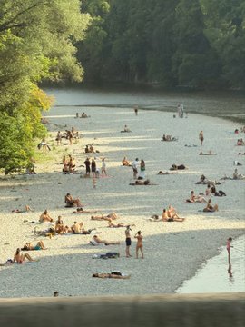A scenic view of people relaxing and swimming at a riverbank or lake shore on a pebbly beach surrounded by lush green trees.