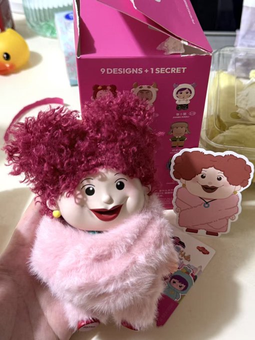 A hand holding a pink plush toy figure with curly magenta hair from a '9 DESIGNS + 1 SECRET' blind box collection, with the corresponding character card and pink packaging box visible in the background.