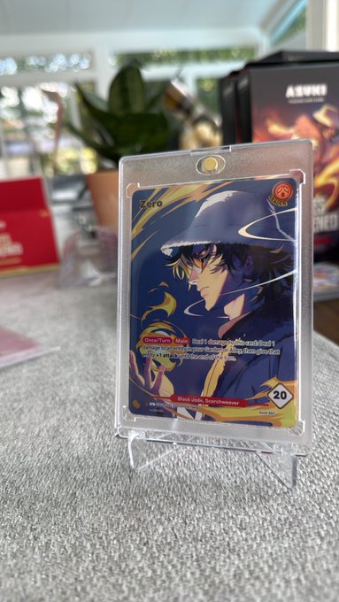 A photograph of a trading card featuring an anime character wearing a dark hat, encased in a plastic sleeve and displayed on a small stand, with 'Zero' and 'Black Hole, Spellbreaker' visible on the card.