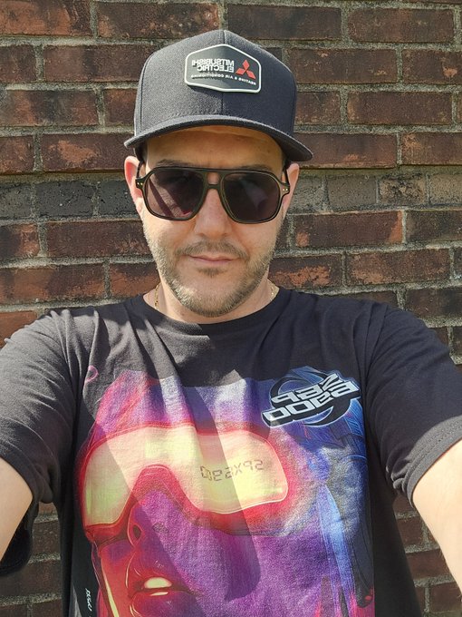 A man wearing a dark cap with a Mitsubishi Motors patch, sunglasses, and a colorful graphic t-shirt poses against a brick wall.