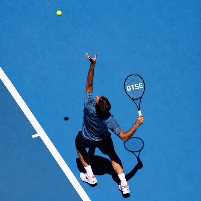 An overhead shot of a male tennis player in a blue shirt serving on a blue court, with 'BTSE' displayed on his racket.