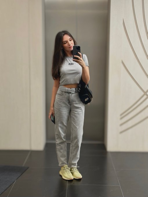 A woman takes a mirror selfie wearing a gray cropped t-shirt, gray jeans, and yellow sneakers, holding a phone and a black Gucci bag.
