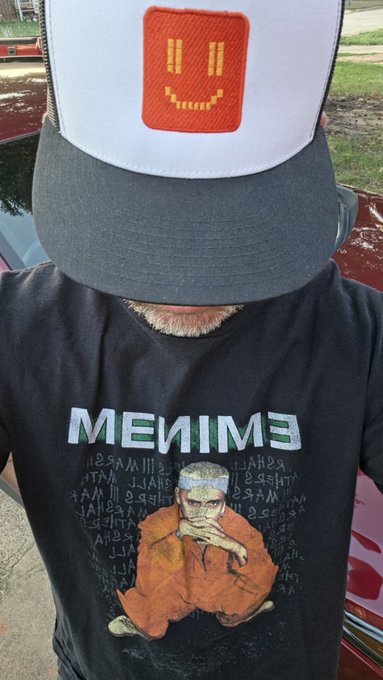 A person wears a trucker hat featuring an orange smiley face patch and a black t-shirt displaying "EINMEИ" text alongside artwork of a meditating figure in orange robes. The post playfully references being "Bullish on $BNKR" and claiming leaderboard rewards, with the image's Bitcoin-inspired orange coin aesthetic (mirrored "EINMEИ" text appears to be a stylized Bitcoin reference) and smiling face reinforcing the crypto/finance meme theme.