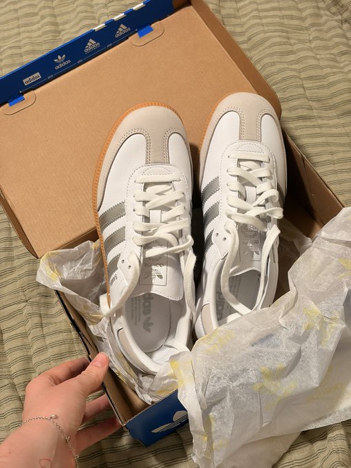 A pair of white Adidas Samba sneakers with gum soles displayed in their original box on a striped fabric.