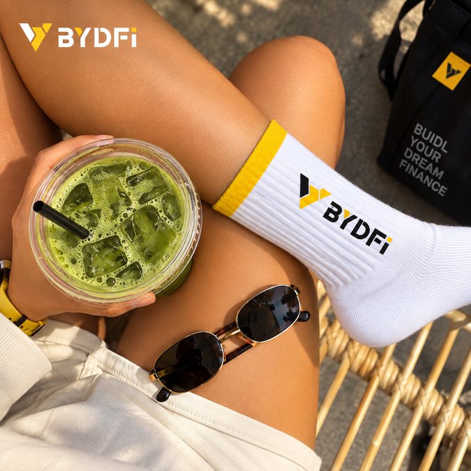 A lifestyle photo showing a person's legs with white 'BYDFi' branded socks, holding a green iced drink, with sunglasses resting on their knee.