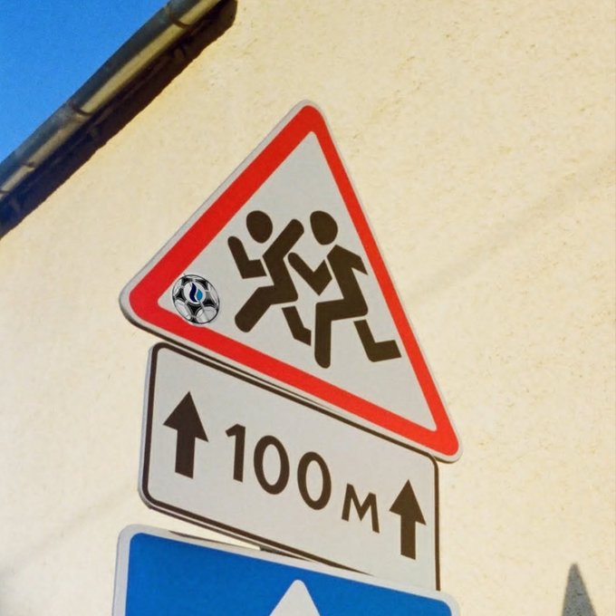 A triangular warning road sign depicts running children, humorously modified with a soccer ball replacing one child's head, positioned above a sign indicating '100 M' in both directions.