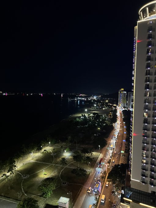 Aerial nighttime view of a coastal city street with traffic and a tall illuminated building on the right.