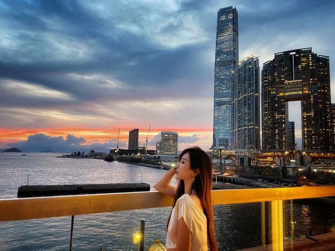 A woman with long dark hair leans on a railing overlooking a harbor at sunset, with illuminated skyscrapers of a city skyline in the background.
