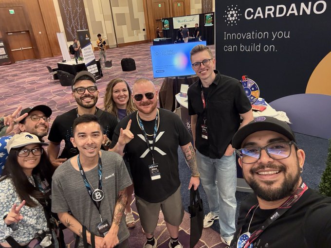 A group of smiling individuals are posing for a selfie at the Cardano booth, which features the slogan "Innovation you can build on." The group's enjoyment aligns perfectly with the caption's declaration of fun.